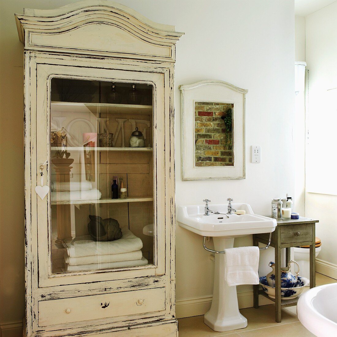 Glass-fronted, vintage-style cabinet and framed, unplastered section of brick masonry above pedestal sink