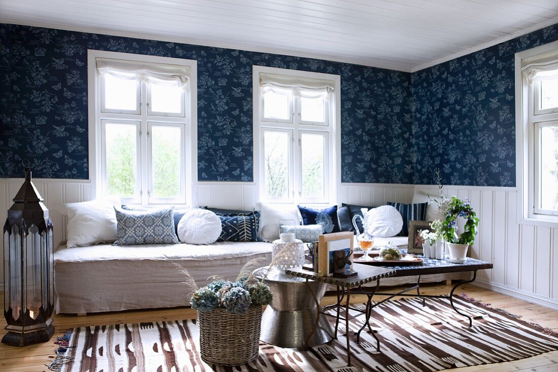 Comfortable living room with various side tables in front of long bench below window and white, half-height wood panelling below blue patterned wallpaper