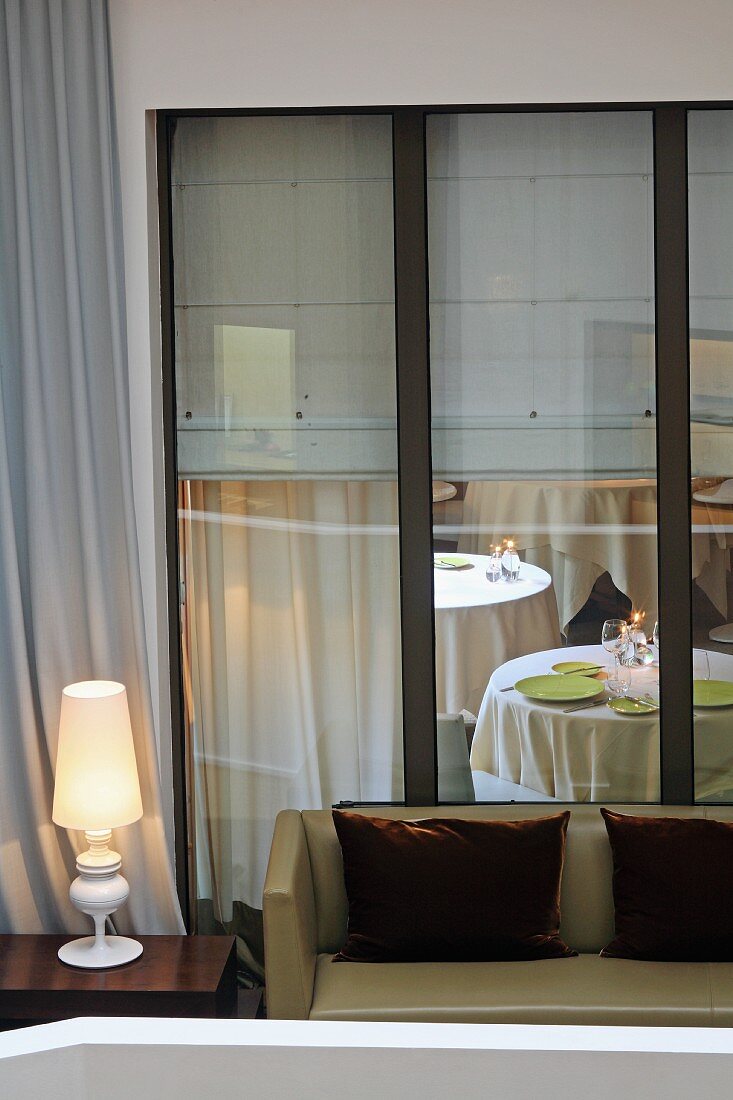 Scatter cushions with brown velvet covers on bench next to lit table lamp on side table in front of tall windows with view of restaurant tables