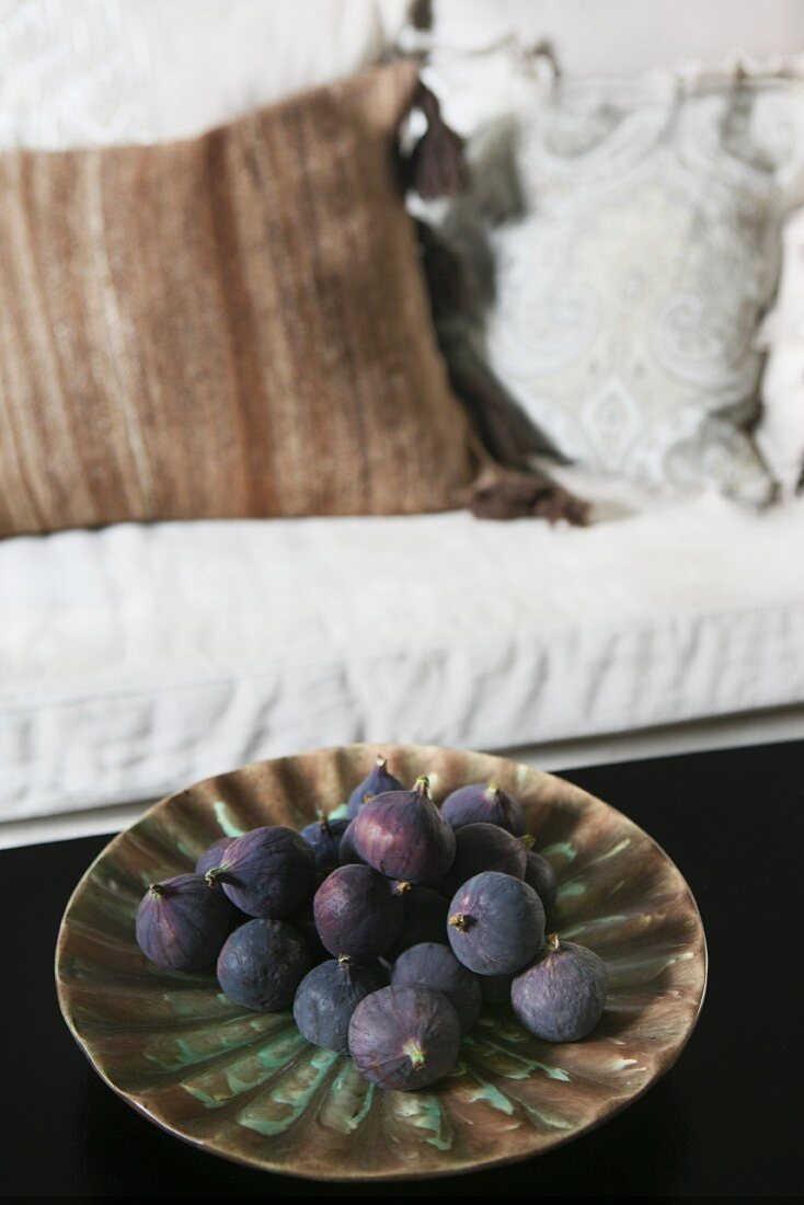 Purple figs on ceramic dish; pale sofa with scatter cushions in background