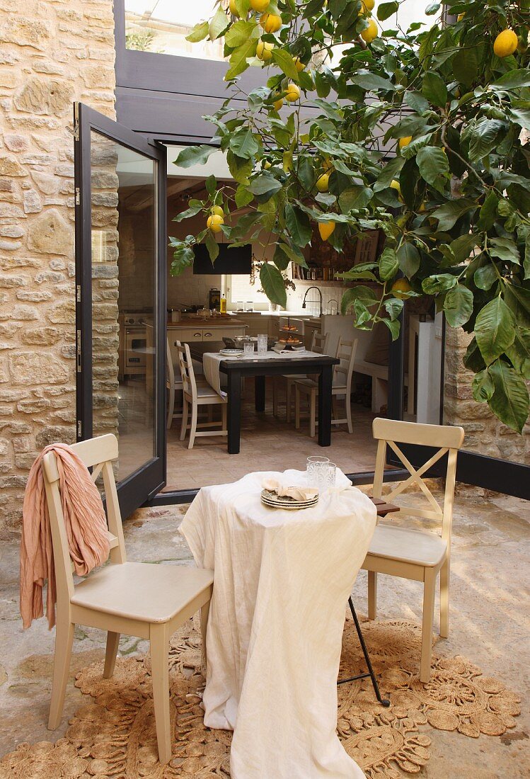 Pale grey kitchen chairs and table with tablecloth on terrace below lemon tree in front of open terrace doors with view of dining area