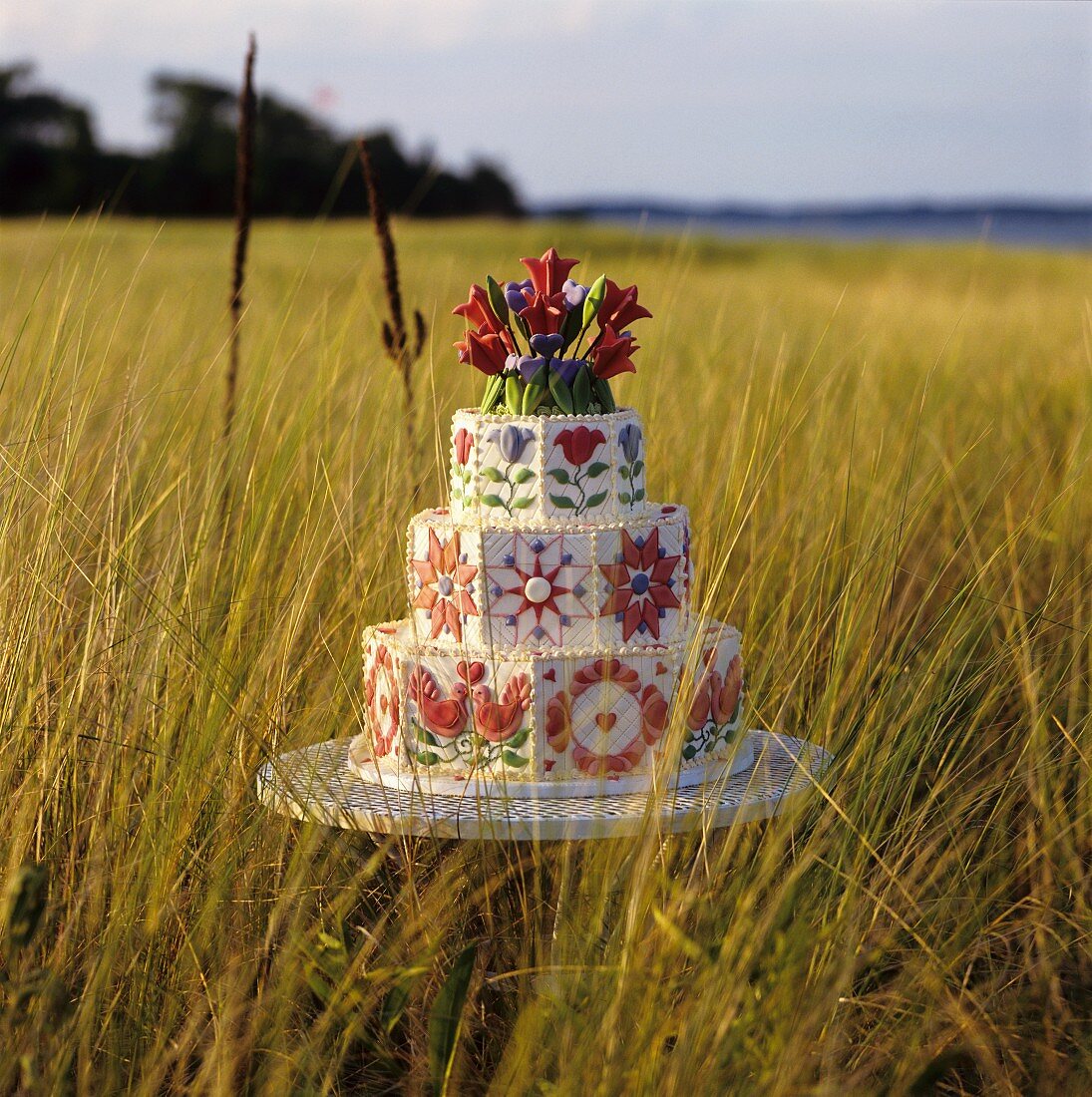 Three Tiered Wedding Cake Decorated with Red and Blue Flowers and Red Love Birds; In a Field