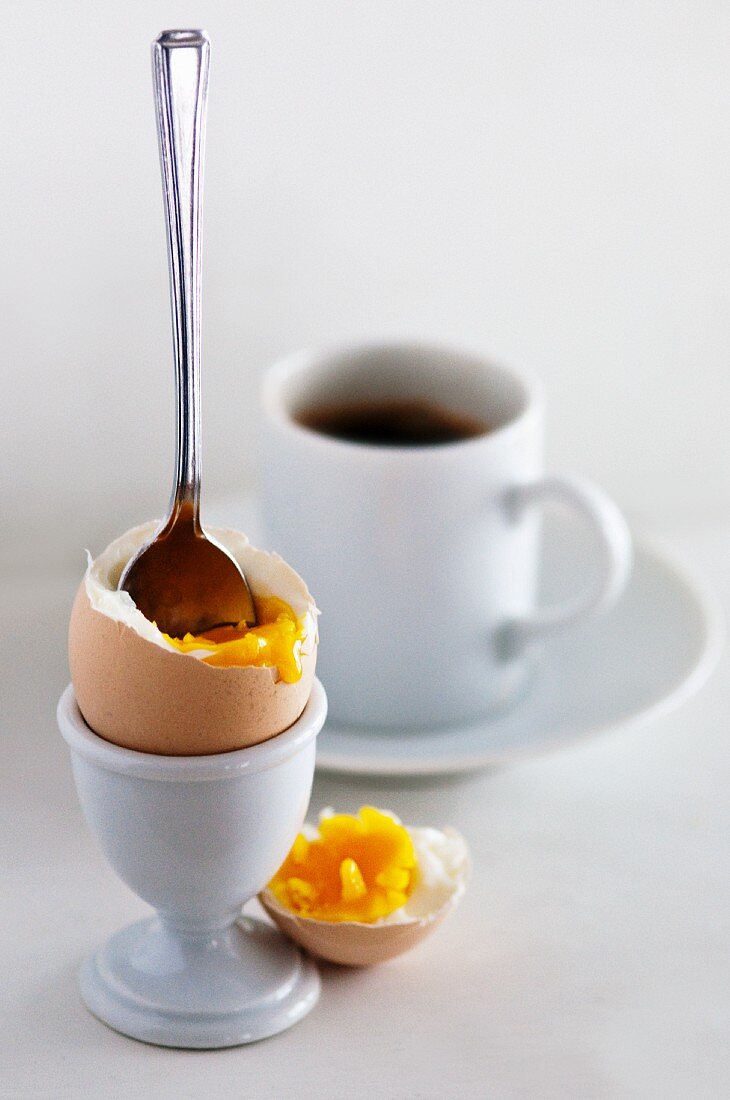A boiled egg with its top cut off, with a spoon in it and a cup of coffee
