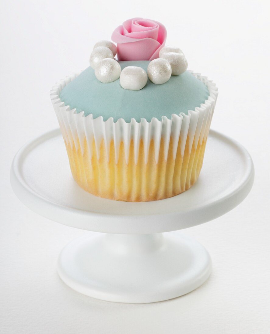 A cupcake decorated with pale blue glaze, sugar pearls and a sugar rose