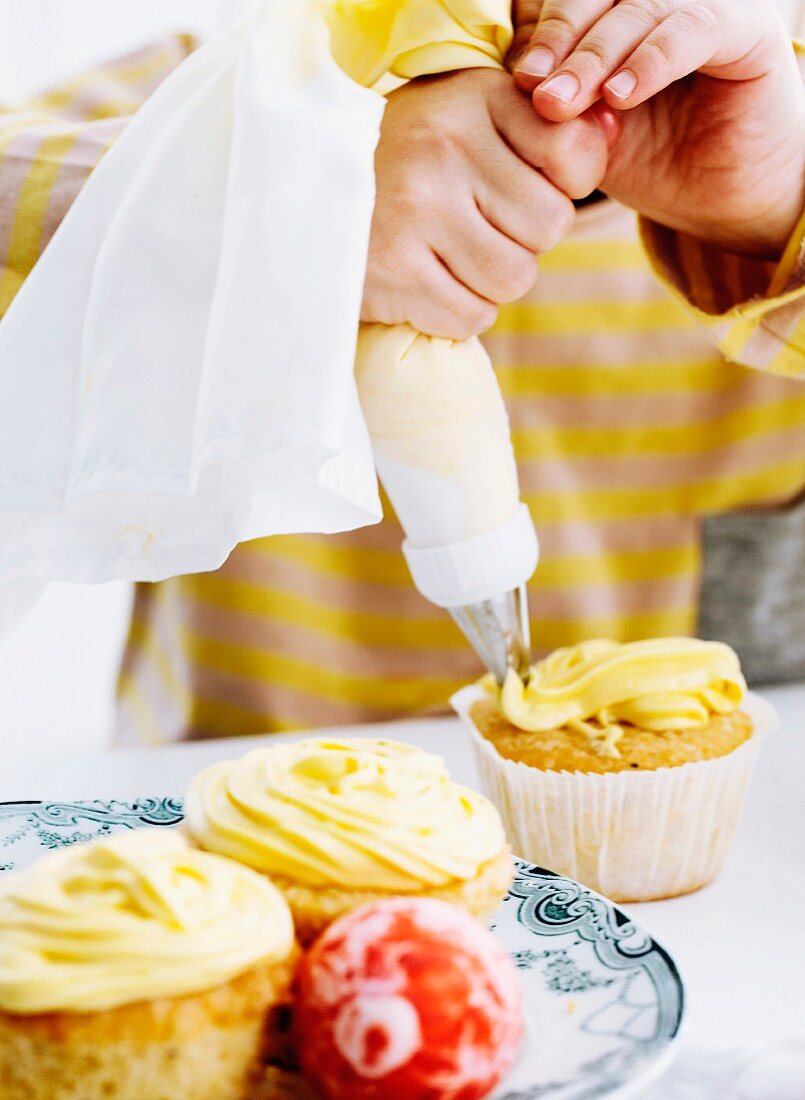 A child's hands using a piping bag full of icing to decorate cupcakes