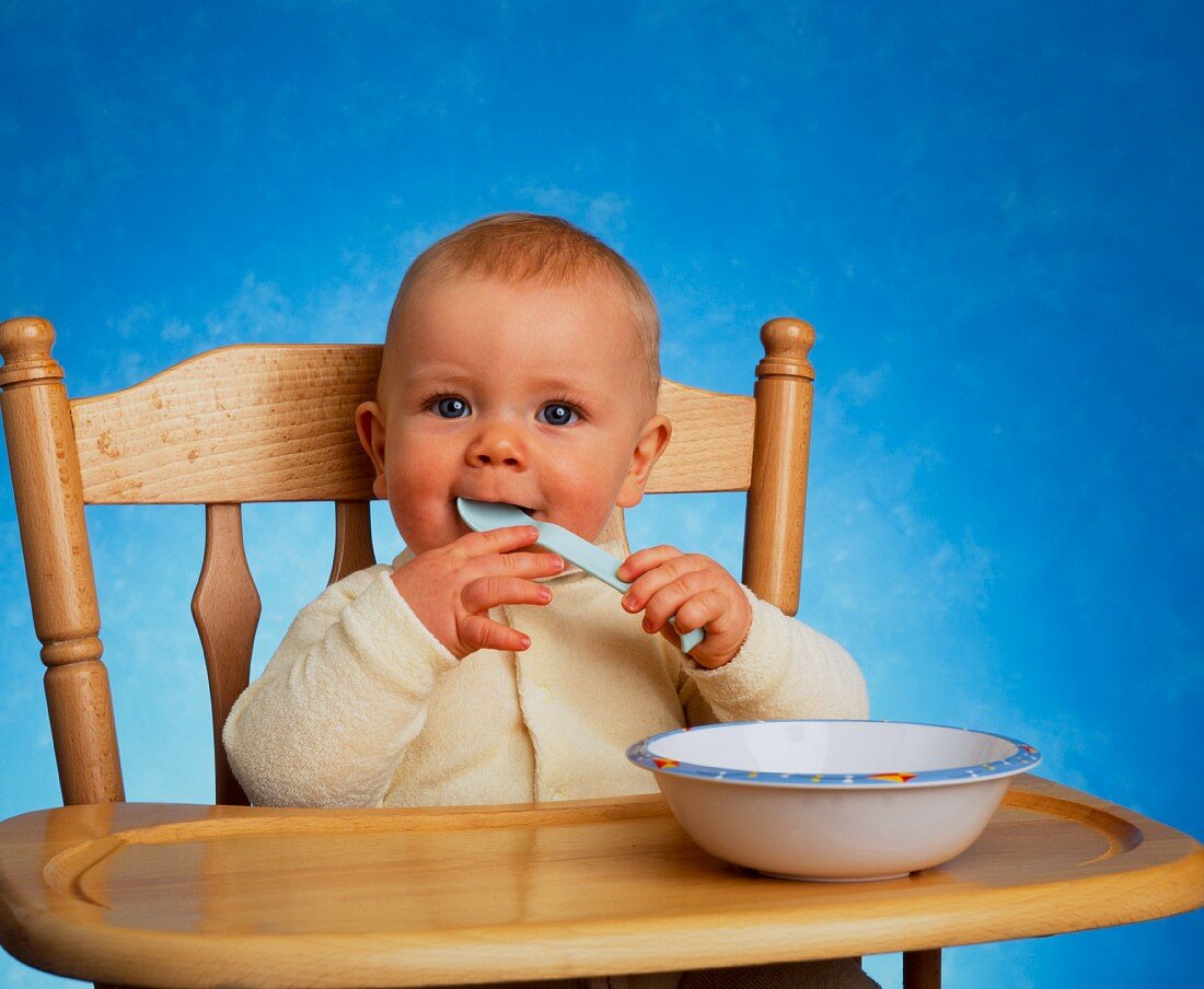 A baby sitting in a high chair with a wooden spoon in its mouth