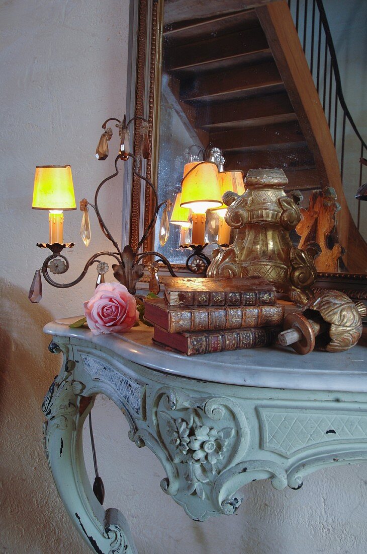 Collection of antique flea-market finds and small lit lamps on Rococo console table; mirror reflecting staircase in background