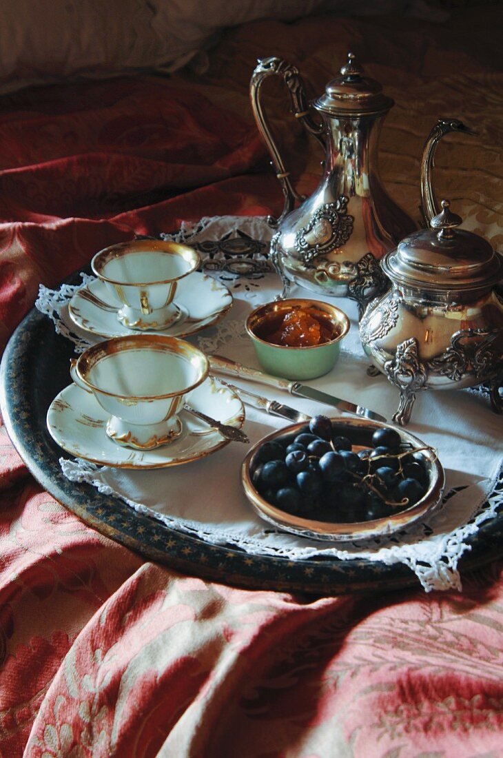 Breakfast tray on bed with silver Rococo pots, gold-rimmed china cups and grapes