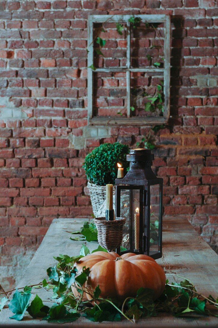 Arrangement of leaves around pumpkin and lit candle in lantern on table in front of rustic brick wall