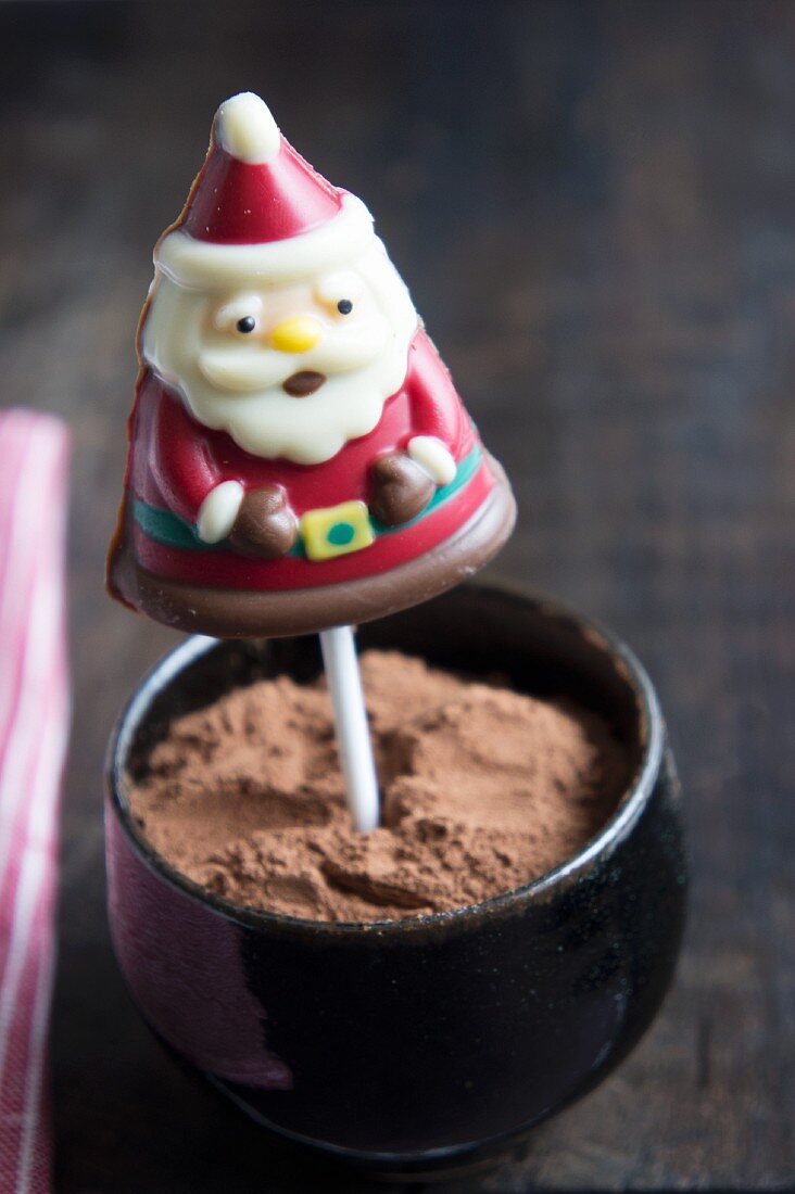 A chocolate Father Christmas on a stick in a bowl of cocoa powder