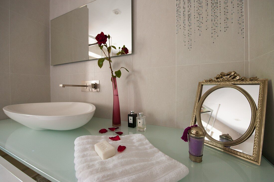 Badezimmerutensilien auf Designerwaschtisch mit Waschschüssel auf Glasplatte und Blumenvase neben Vintage Spiegel