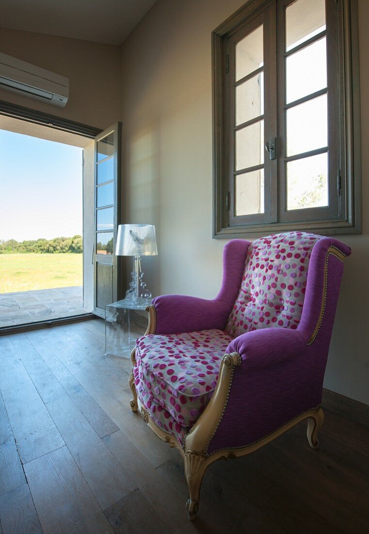 Rococo reading chair with colourful, spotted designer upholstery in minimalist room with traditional ambiance and view of landscape through open garden door