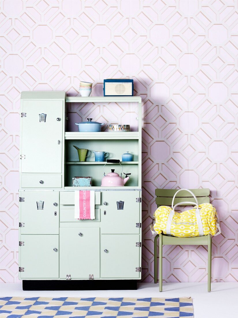 Pastellgrüner Küchenschrank im Fiftiesstil neben Stuhl mit Tasche vor Wand mit dreidimensionalem Muster auf Tapete