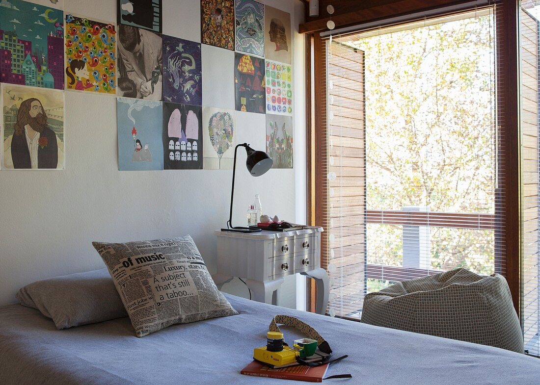 Single bed below posters pinned on wall with vintage bedside table and open louver blinds on balcony door to one side