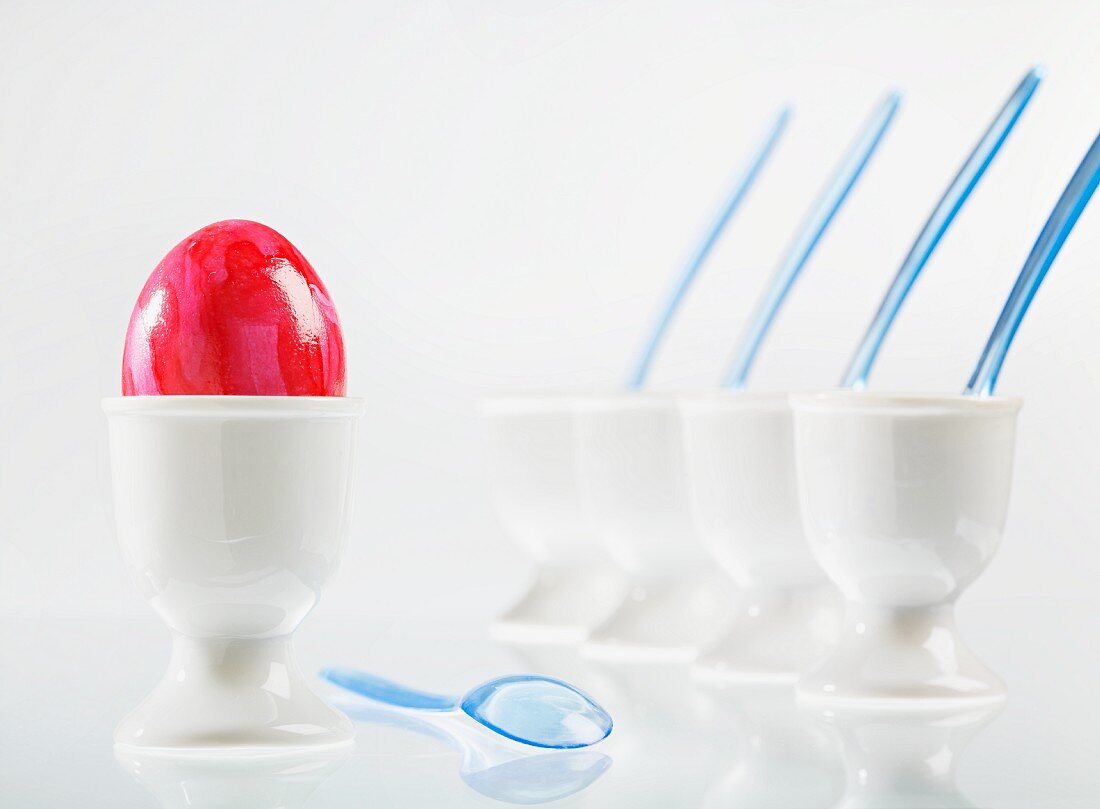 An egg, coloured red for Easter, next to a row of white eggcups
