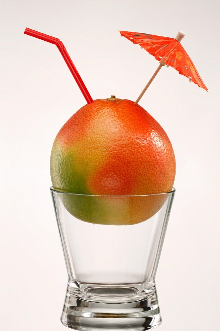 A grapefruit on a glass with a cocktail umbrella and a drinking straw