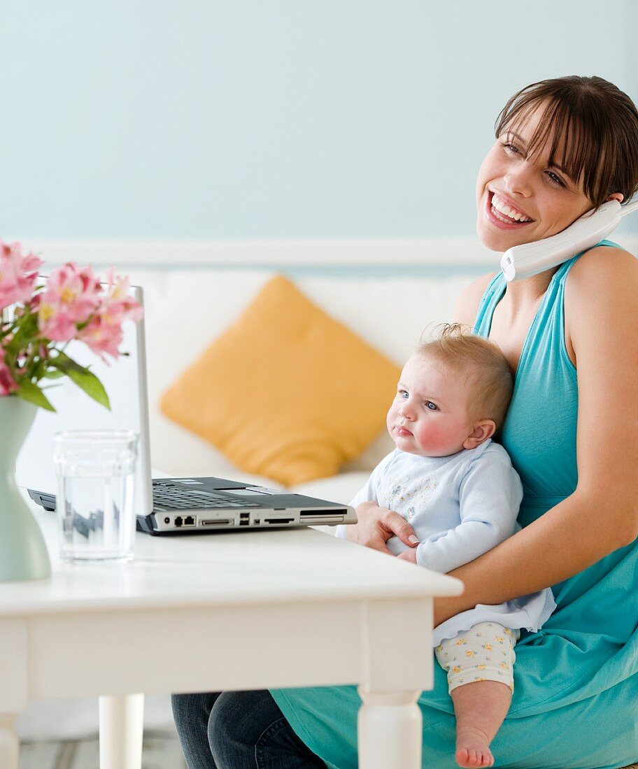 Mutter mit Baby und Telefon vor einem Laptop