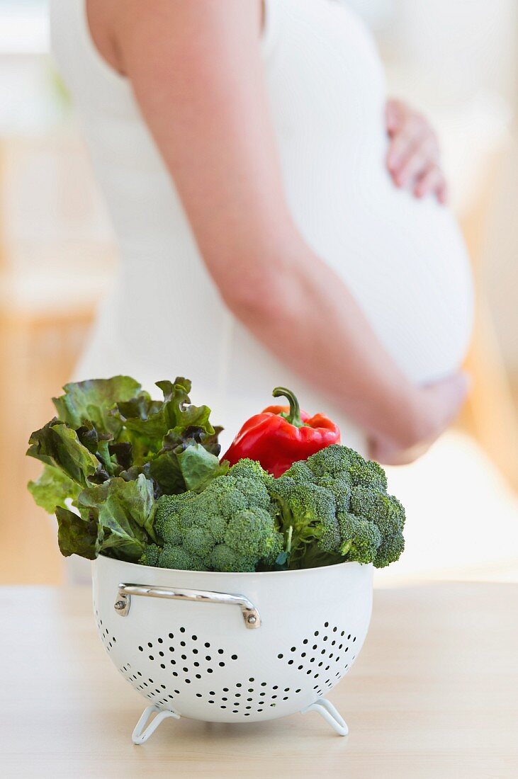 Fresh vegetables in colander, pregnant woman in background