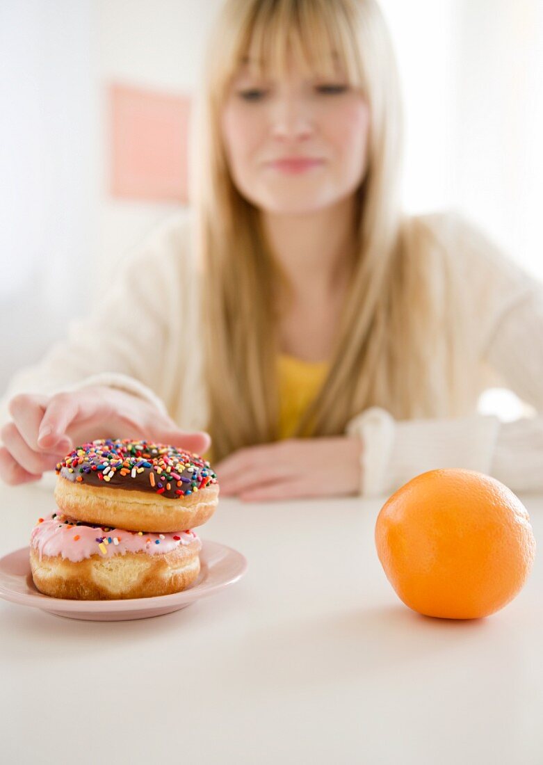 Junge Frau sitzt am Tisch vor Donuts & einer Orange