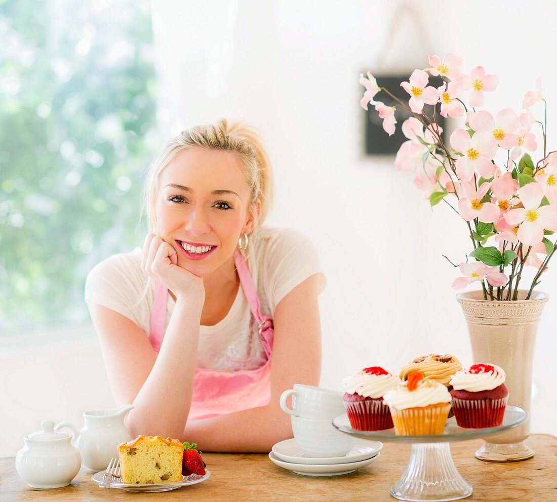 Smiling young woman in apron leaning on table with cakes