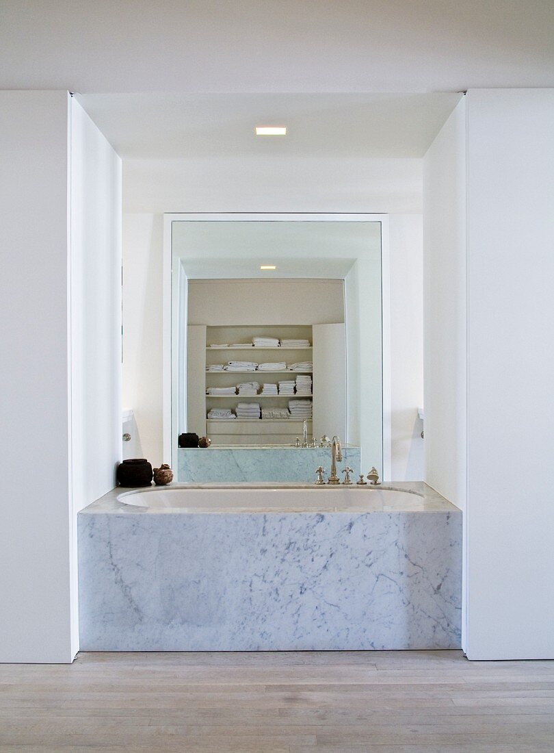 Bathtub with marble sides built into open niche and floor-to-ceiling mirror on opposite wall