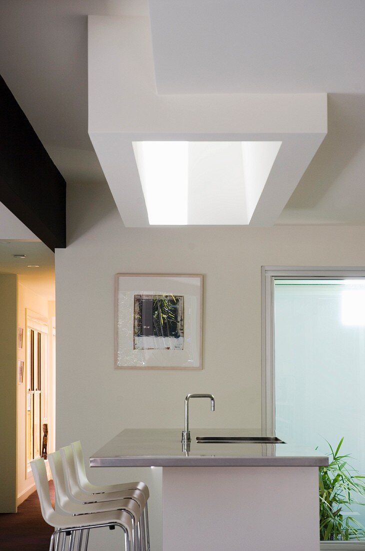 Schachtförmiges Oberlicht über Kücheninsel mit Edelstahlblech Verkleidung und einer Reihe weisser Barhocker