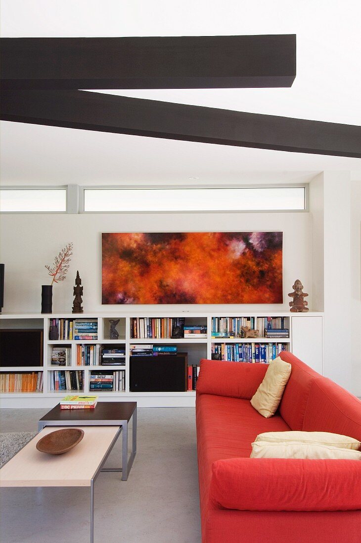 Red sofa under dark ceiling elements in a modern, bright living room; paintings and ethnic art on bookshelves in the background