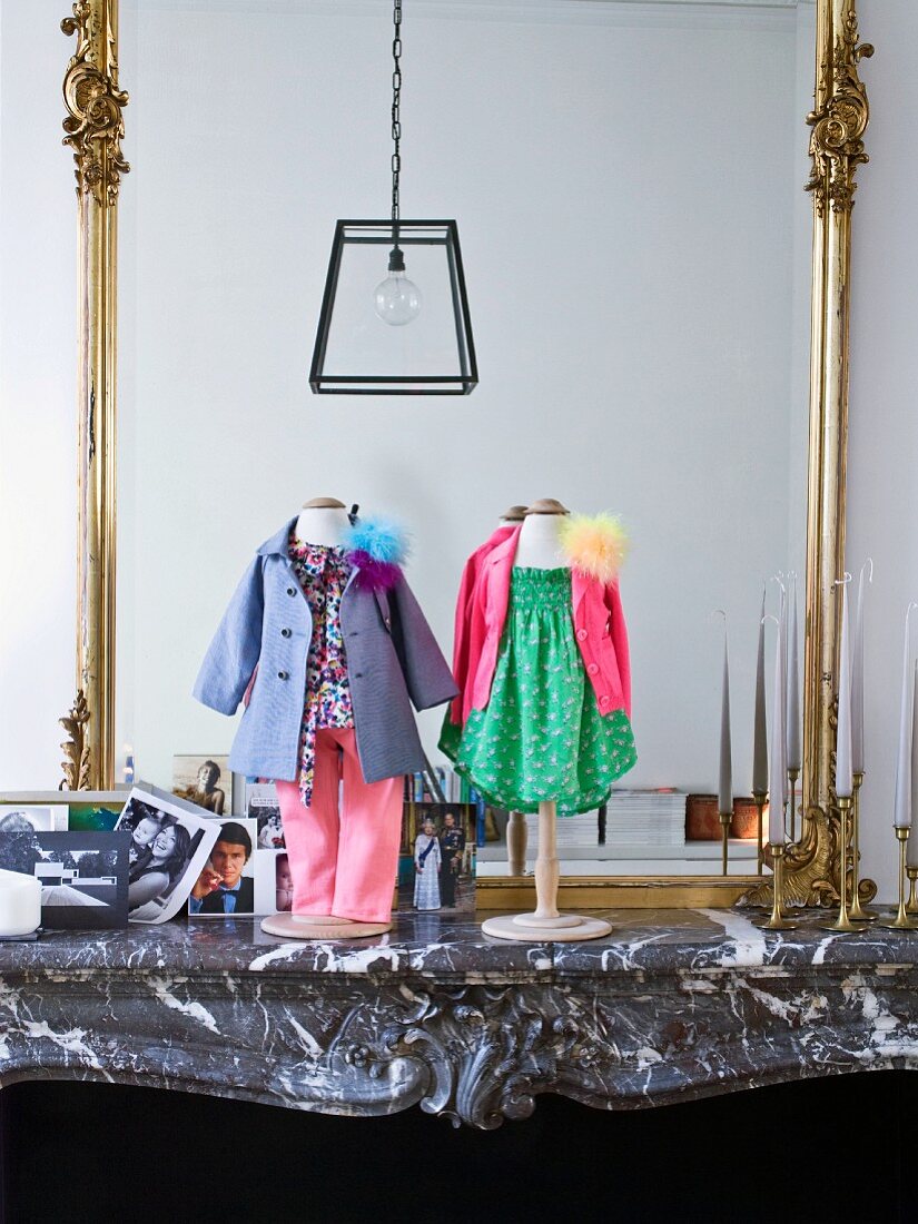 Dolls' clothes on stands on stone mantelpiece in front of gilt-framed mirror