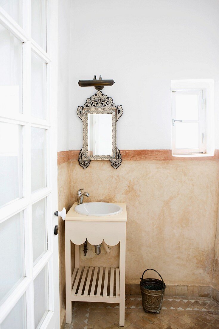 Moroccan bathroom in soft earthy shades with ornate mirror, wooden washstand, half-height marbled wall and terracotta floor