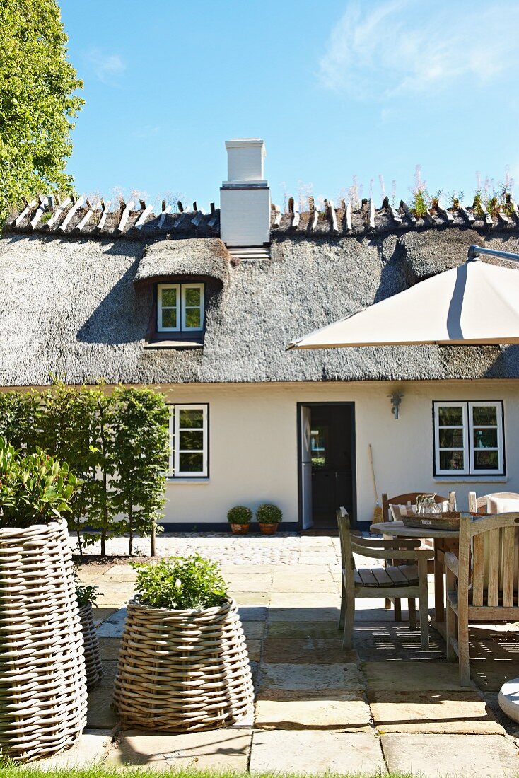 Seating area on terrace with wicker planters, armchairs and table in front of traditional thatched house