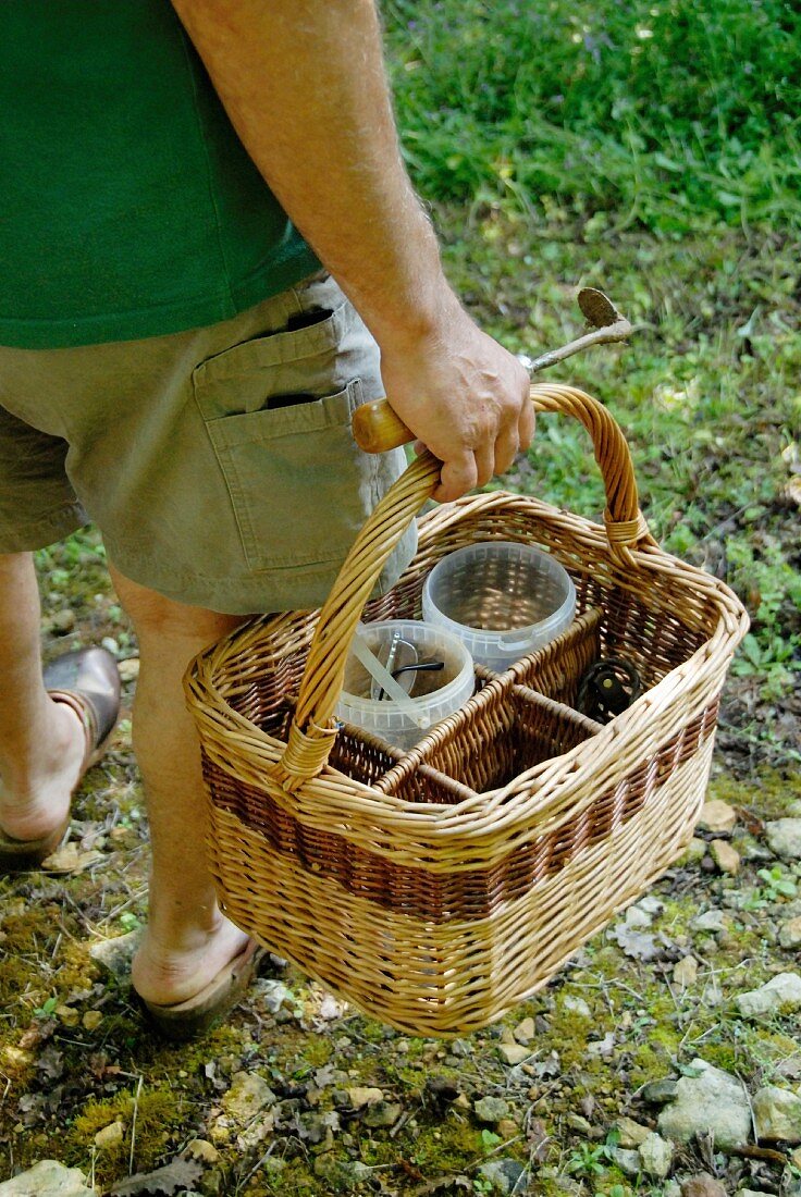 A man standing on a loose stone path in a field holding a small gardening tool and a bottle carrier in his hand