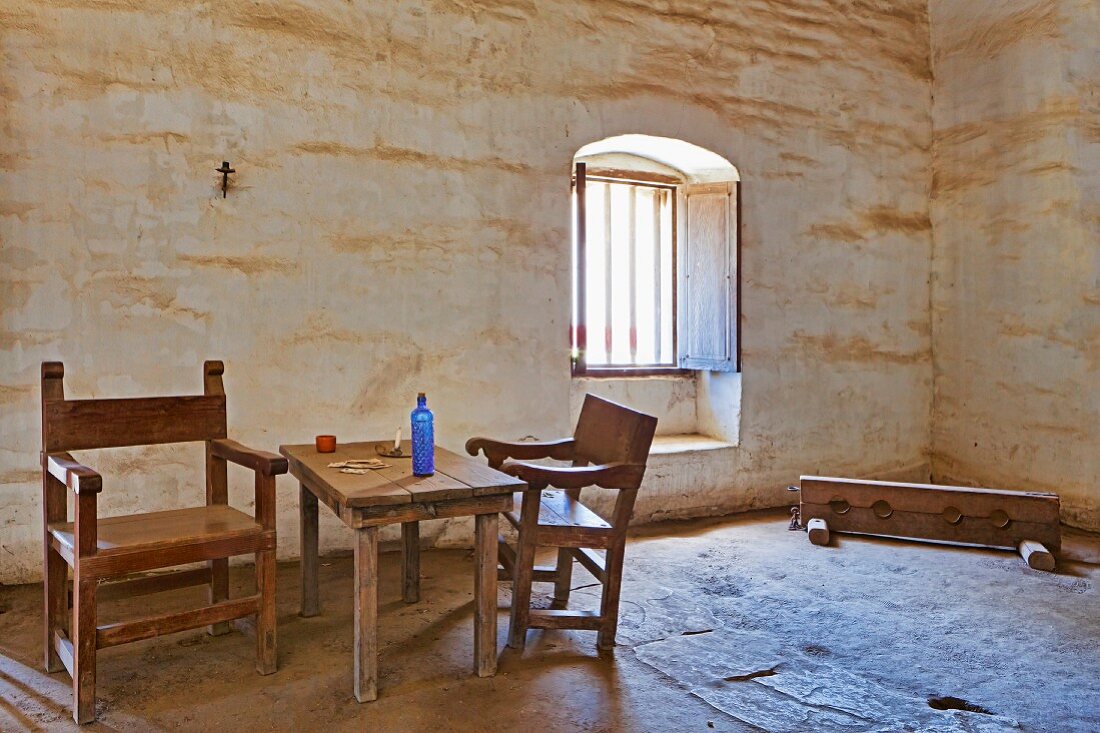 Holztisch mit Stühlen und eine umgefallene Bank in einem Raum mit Lehmziegelwänden, Mission La Purisima State Historic Park, Lompoc, Kalifornien