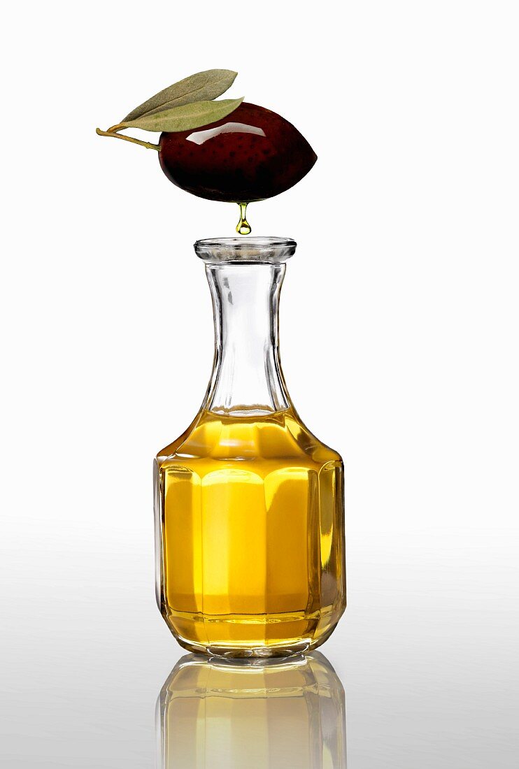 A black olive with an oil droplet over a carafe of olive oil