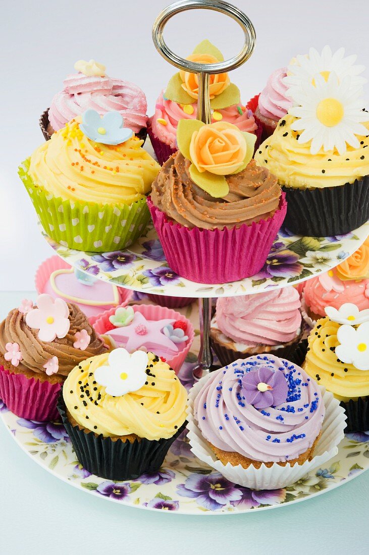 close up of a selction of iced purple, yellow and pink cup cakes decorated with icing flowers on a purple pansy flowered cake stand and a white background