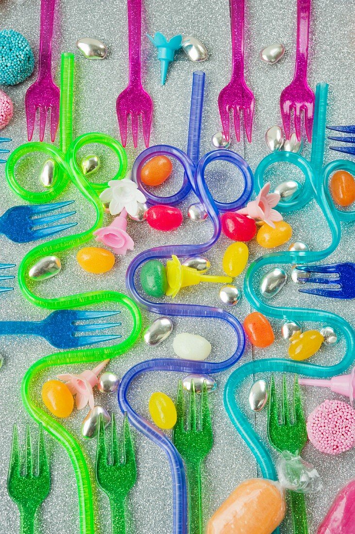 graphic overview of coloured plastic party forks and blue and purple curved drinking straws on a silver glittery background with sweets