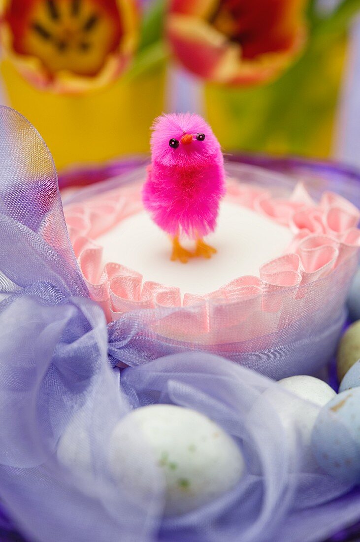 close up of single pink chick on top of a square easter cake tied with a purple chiffon bow surrounded by chocolate eggs and red and yellow tulip flowers