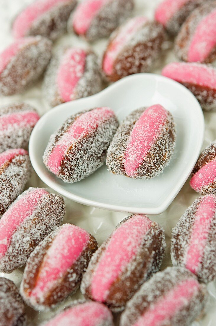 close up of pink marzipan stuffed dates with two laying in a white heart shaped ceramic dish