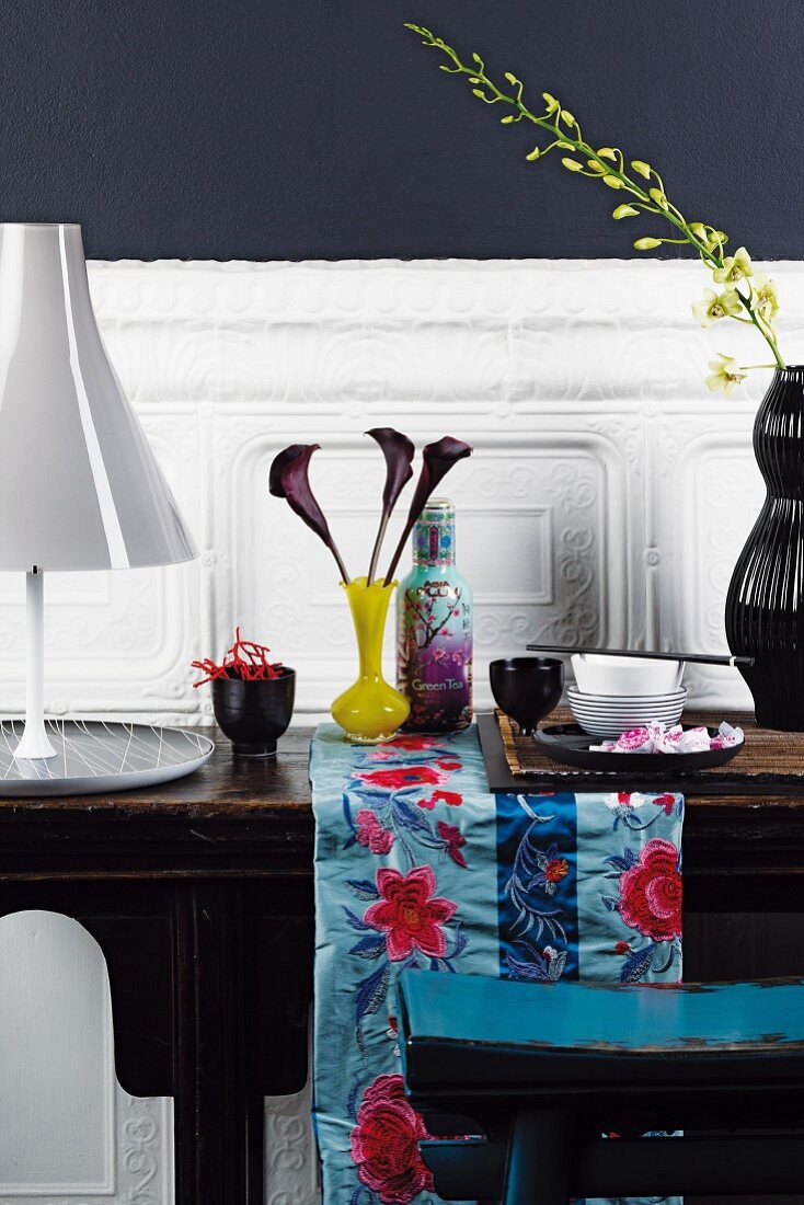 Antiker Holztisch an weisser Wandverkleidung; auf dem Tisch asiatisches Geschirr, Vasen und eine elegante Tischlampe in Grau mit ausladendem Lampenfuss