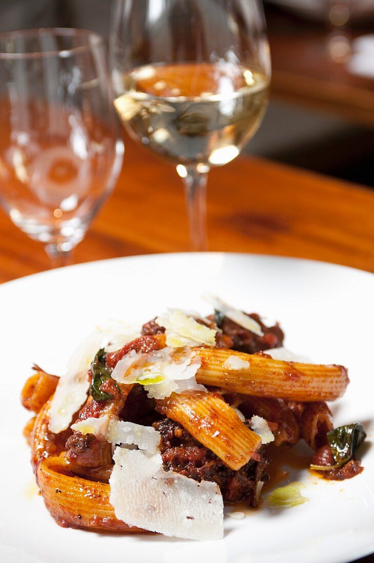 Rigatoni with Short Ribs and Shaved Parmesan Cheese; On a White Plate; Glass of White Wine in Background