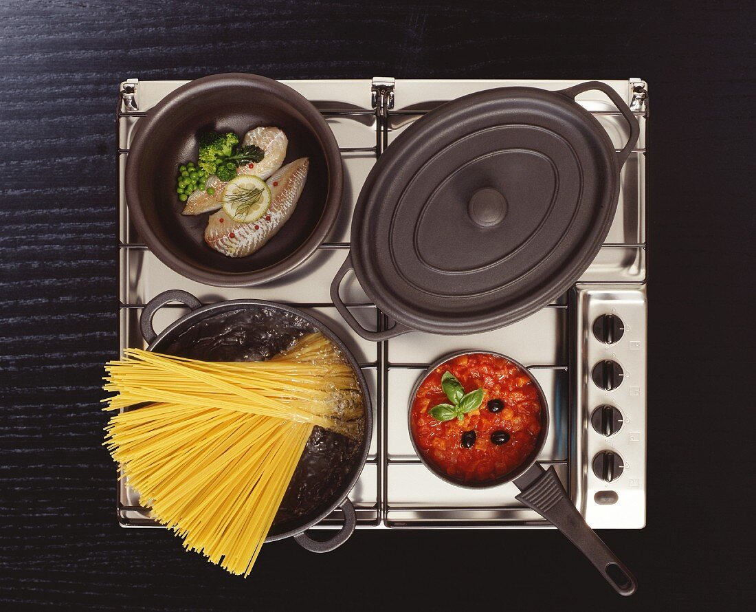 Edelstahlherd mit kochendem Nudelwasser, Fisch in der Pfanne, Tomatensauce und Schmortopf