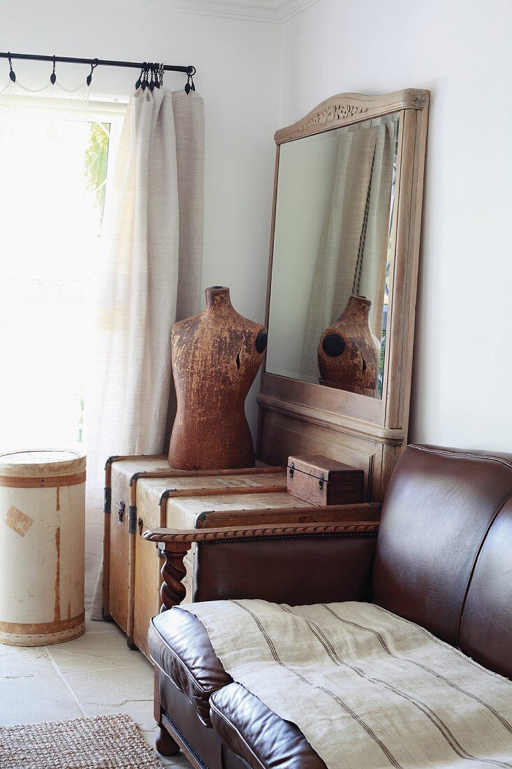 Traditional, brown leather sofa next to wooden trunk and tailor's dummy in front of framed mirror in corner of room next to window