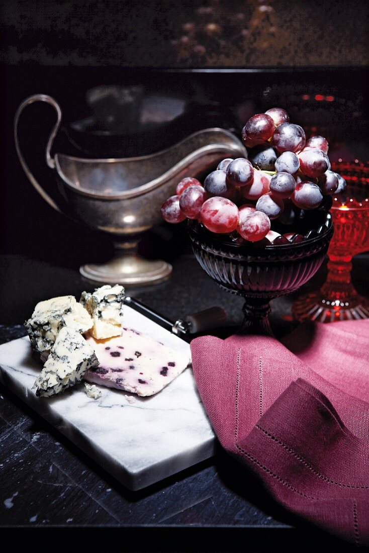 Blue cheese on a cheeseboard, a bowl of red grapes and an antique sauce boat