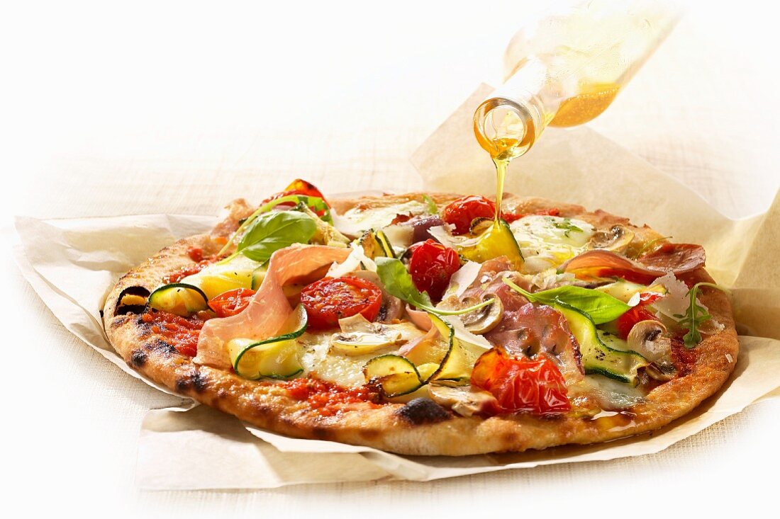 Olive oil being drizzled over a ham and vegetable pizza