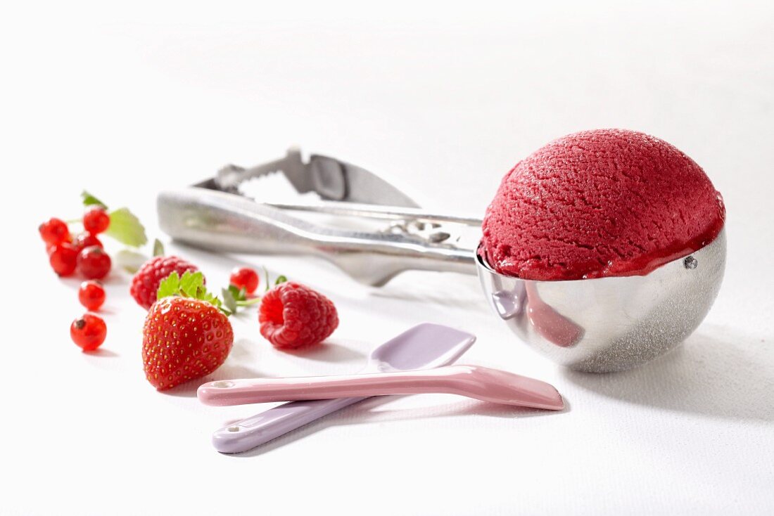 A scoop of raspberry ice cream in an ice cream scoop, with fresh berries and ice cream spoons