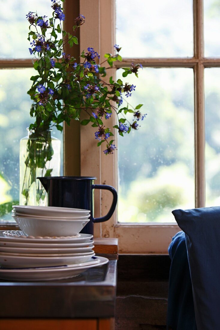 Vintage Tellerstapel und Vase mit blauen Blumen am Fenster