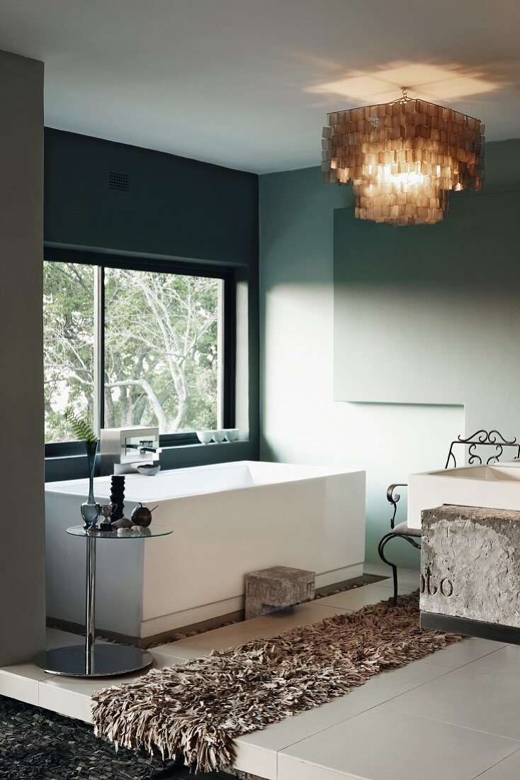 Designer-Badezimmer - Flokatiartiger Läufer auf weißem Fliesenboden vor Badewanne am Fenster und Deckenleuchte aus hellbraunen Perlmuttplättchen