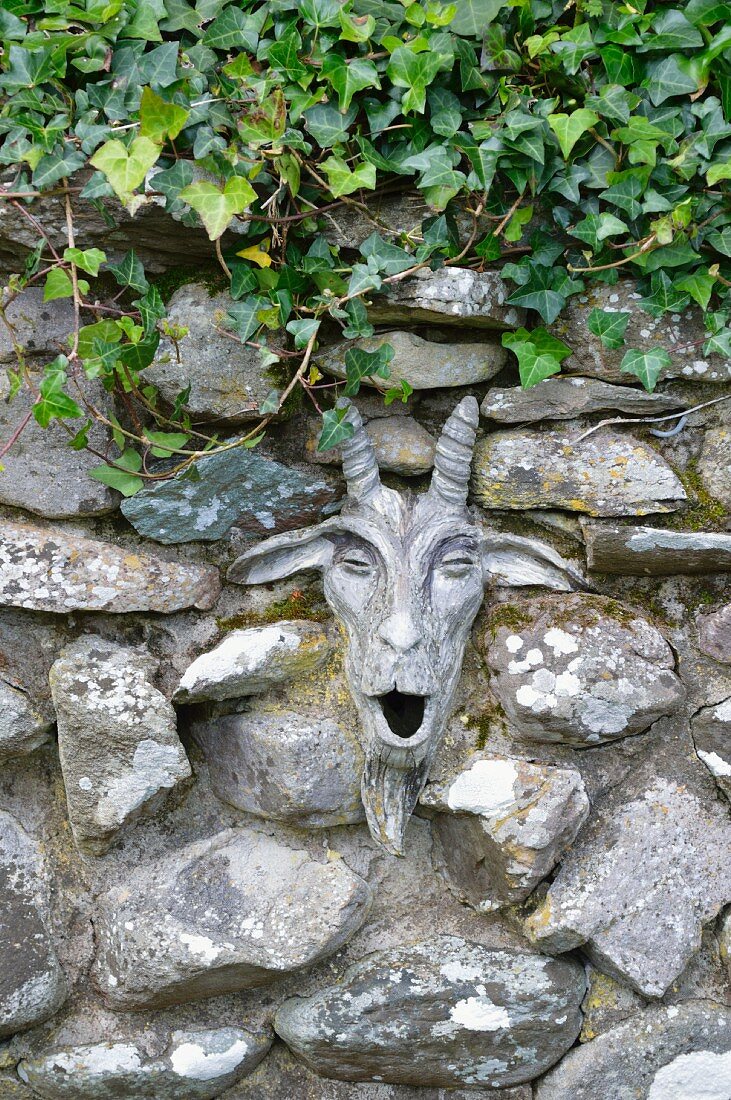 Natursteinmauer mit Efeu und origineller Ziegenkopf-Skulptur als Deko im Cottagegarten