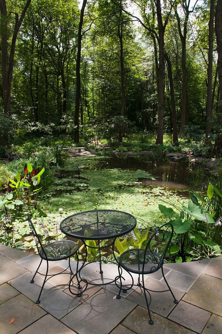 Gartentisch mit Stühlen aus Metallgeflecht auf Terrassenboden aus Steinfliesen und Blick auf Teich in ländlichem Garten
