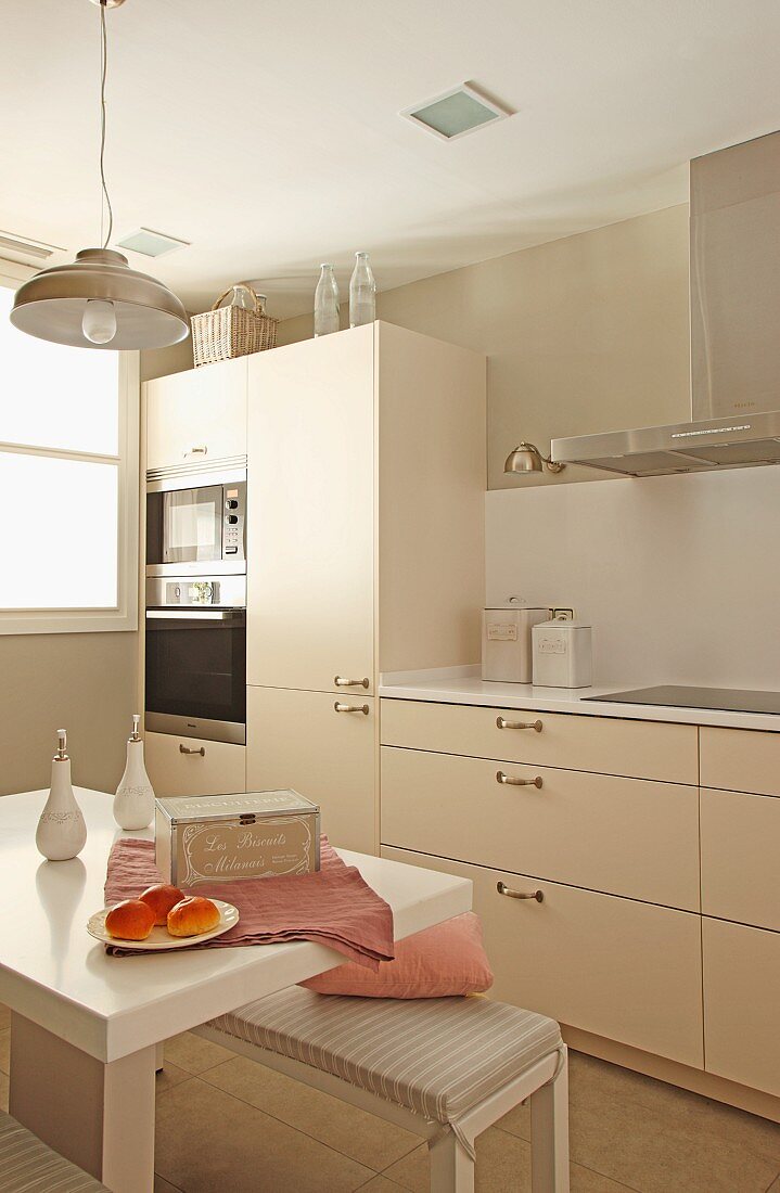 Pale beige, contemporary fitted kitchen and small, white breakfast table and upholstered bench