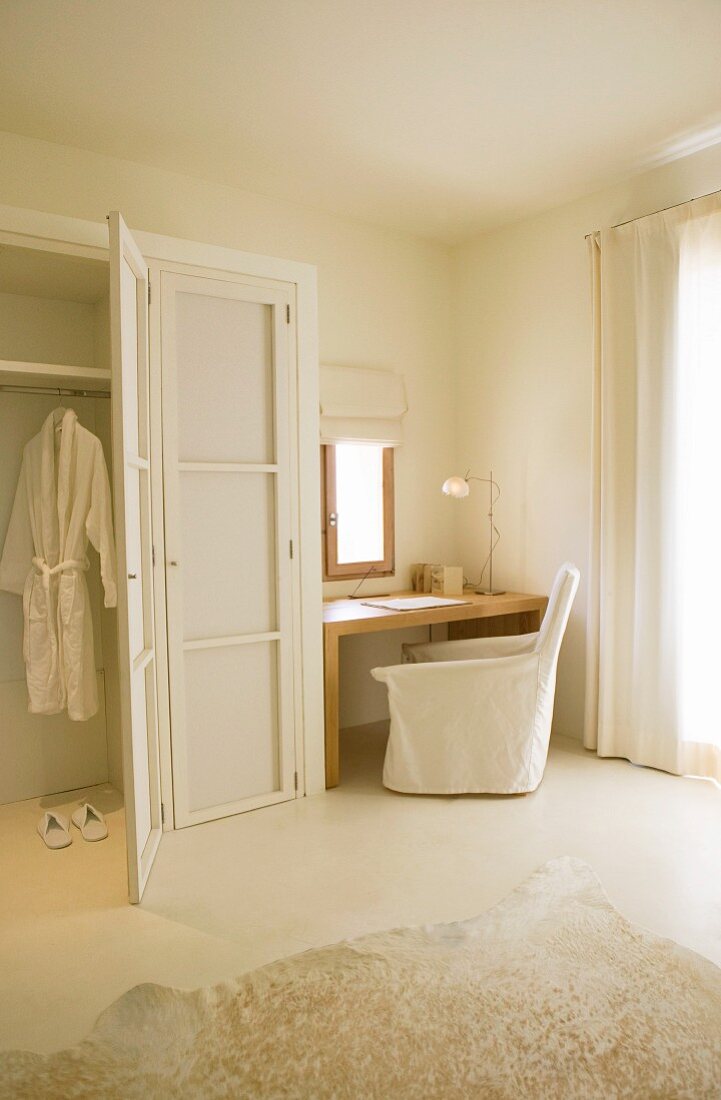 Minimalist simplicity in white bedroom with loose-covered armchair and desk below small window